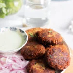 a plate of shami kabab with onions, a green raita/chutney and cilantro in a jar in the background